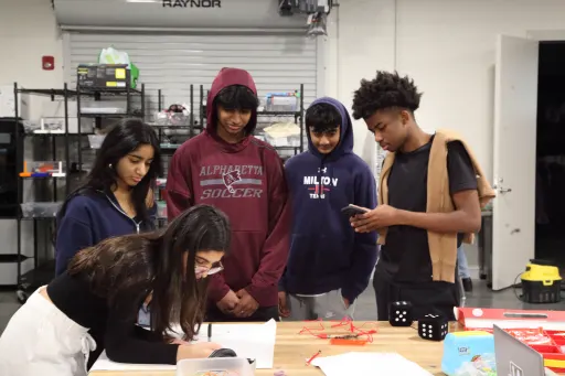 Students draft idea on wide paper on-top of a wooden workbench inside of a industrial looking room with a large garage door behind them, with shelves infront of the garage door.